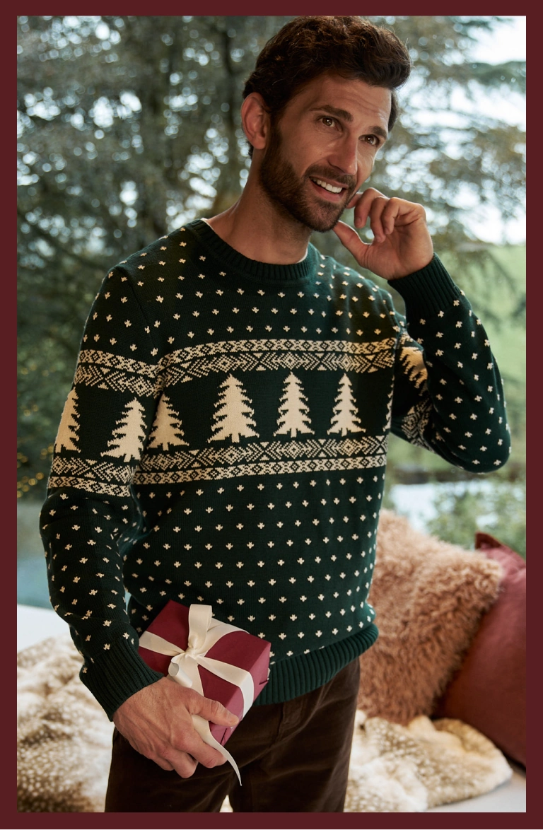 Homme portant un pull de Noël vert foncé à motif sapin et tenant un cadeau, avec un visuel indiquant 50 % de réduction